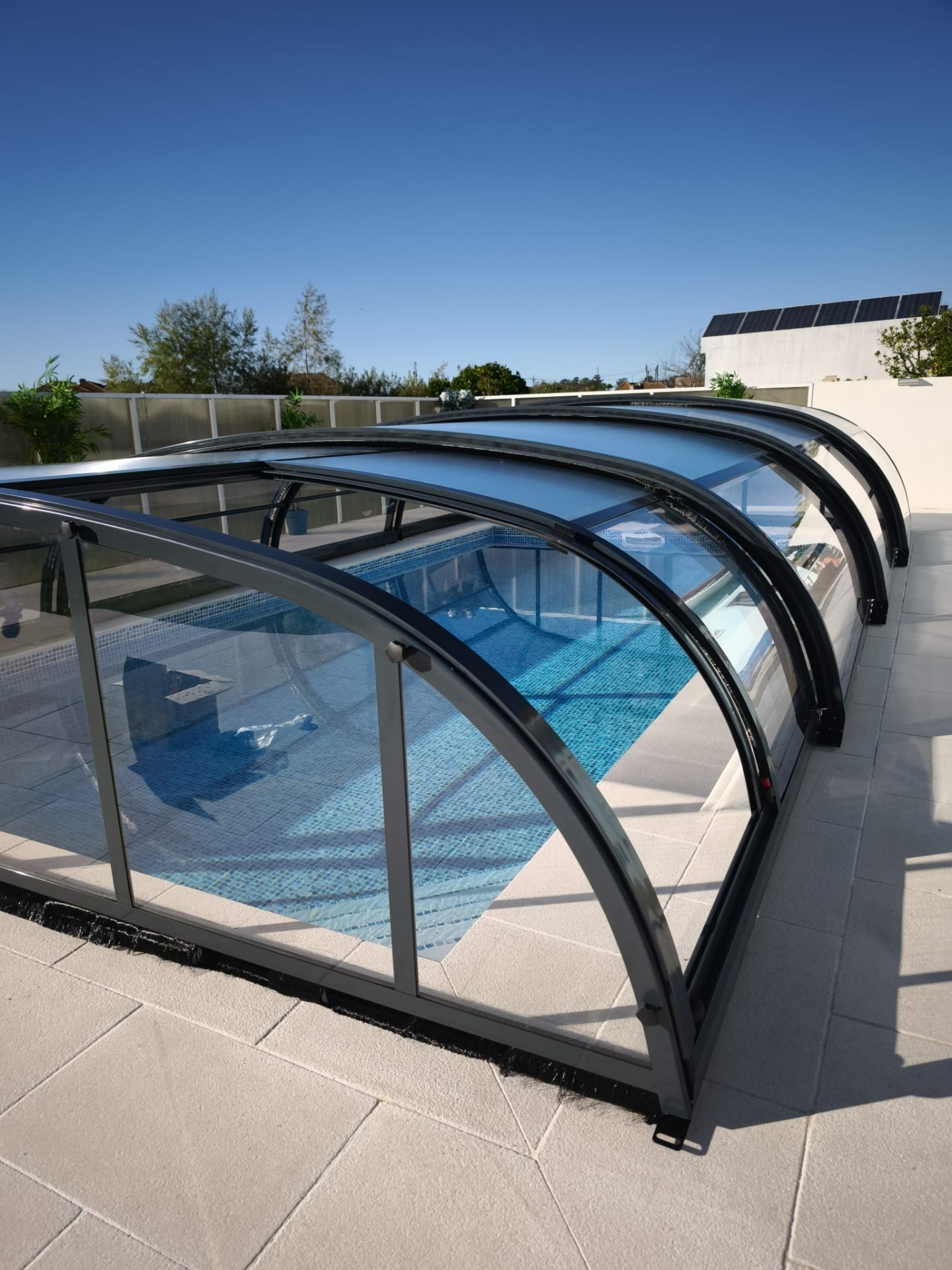 Cobertura de piscina com estrutura de vidro e metal preto, vista do terraço ao céu azul.
